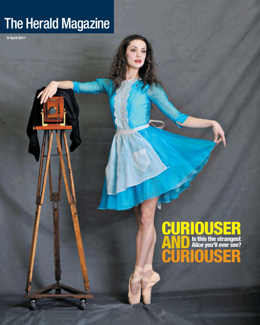 Principal ballet dancer dressed at Alice from Scottish Ballet production of Lewis Carol's much loved story Alice Adventures in Wonderland for Herald Magazine cover story with portraits of cast. Portrait of dancer with dark hair wearing blue dress and large format vintage camera on tripod.